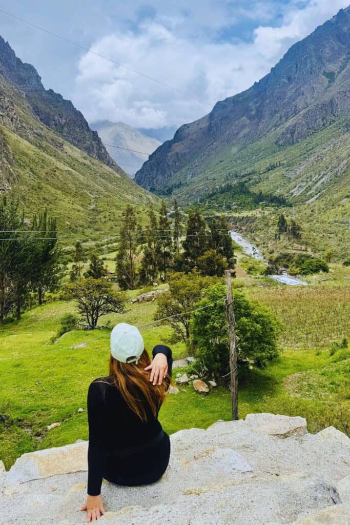Frau sitzt auf Stein und bewundert die grüne Täler- und Bergwelt der Anden in Peru mit Fluss und Wolken.