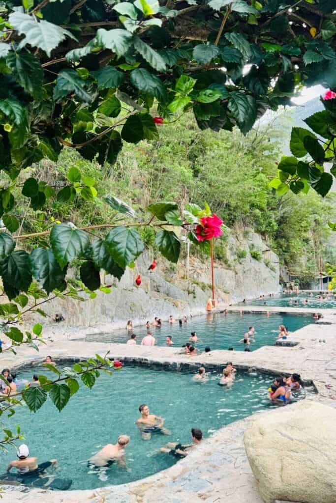 Alt: Thermen mit Menschen im warmen Wasser, umgeben von Natur und grünen Blättern.